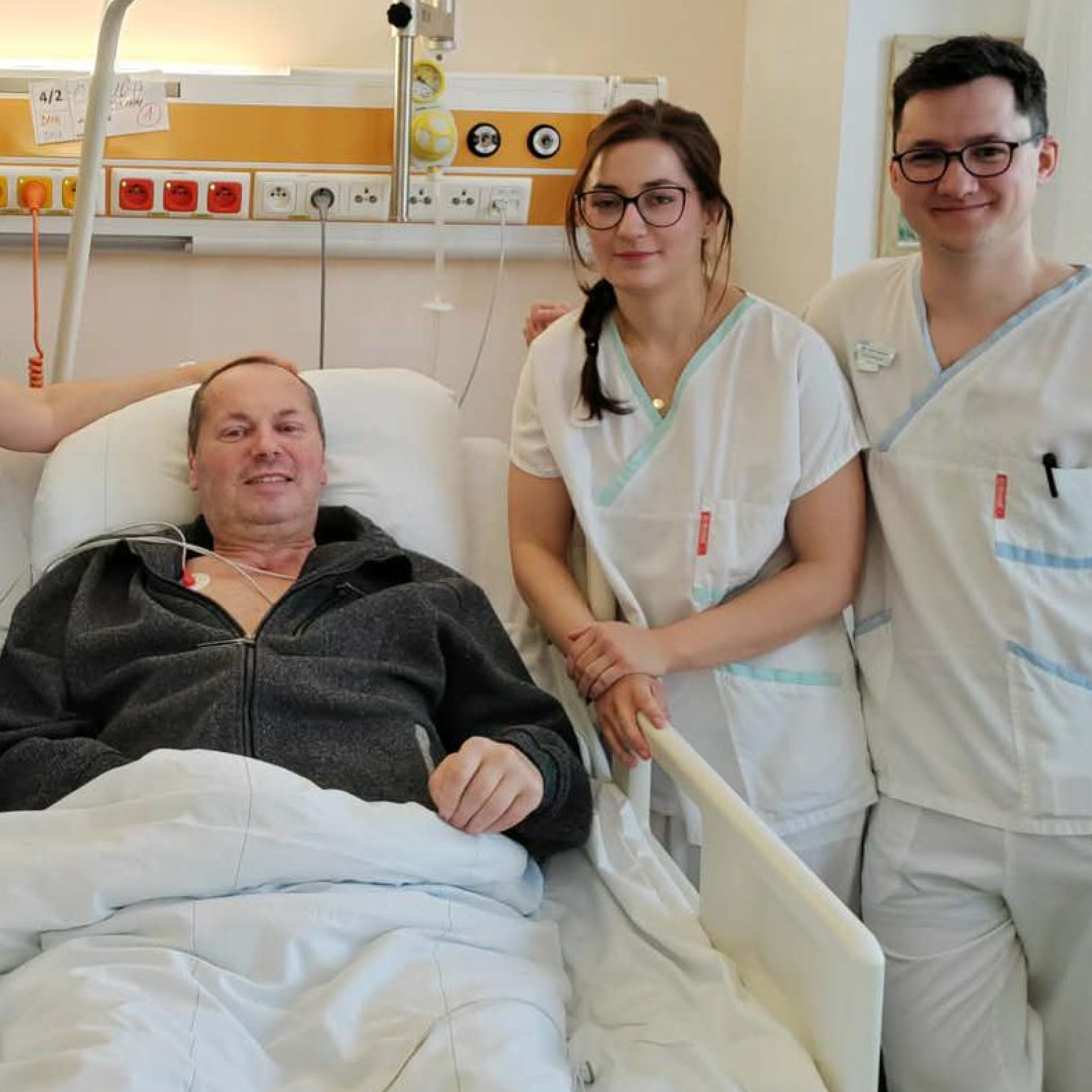 Pacient na lůžku a dvě osoby v bílém zdravotnickém oblečení.