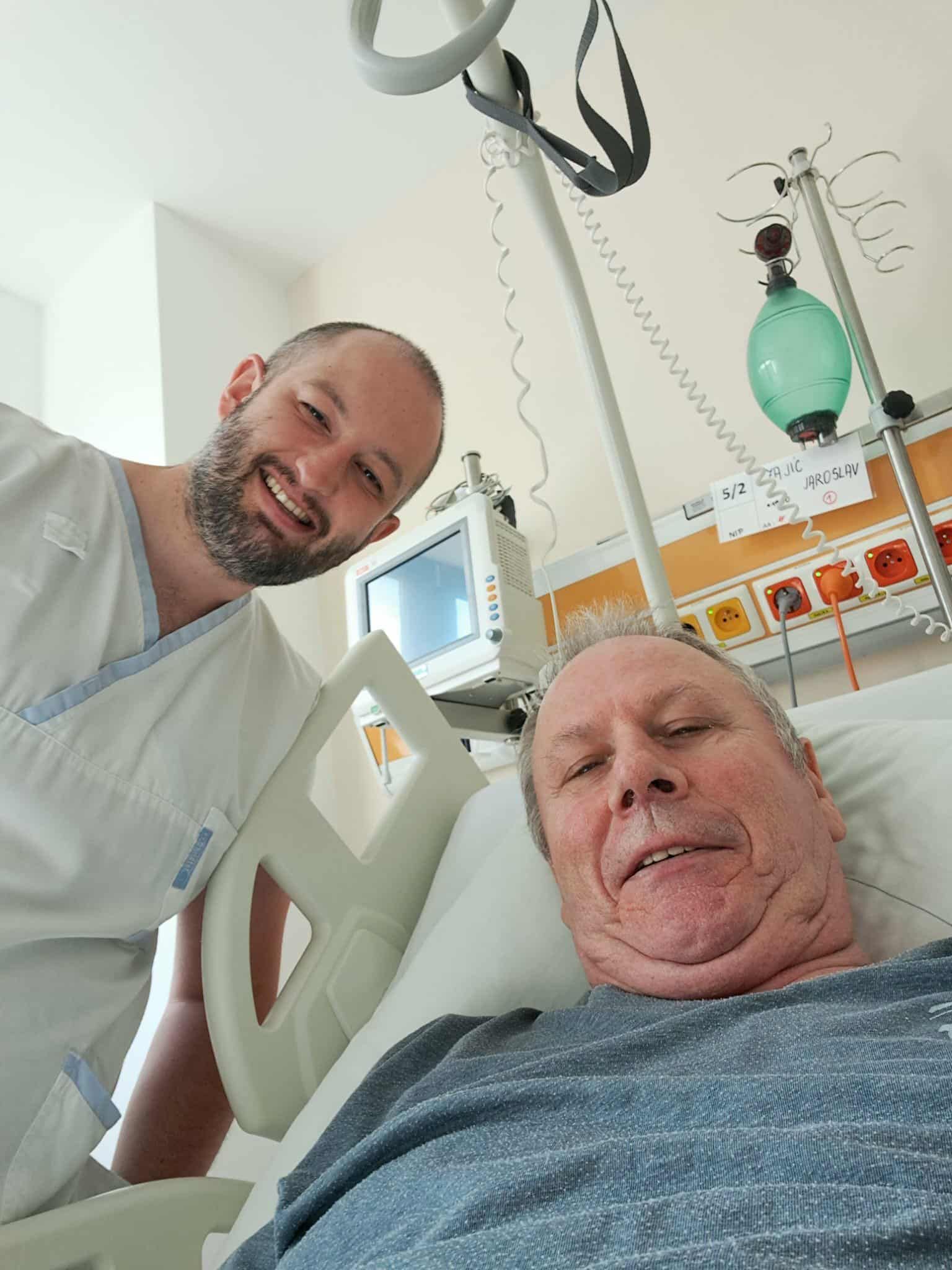 Muž v bílém oděvu vedle pacienta na nemocničním lůžku